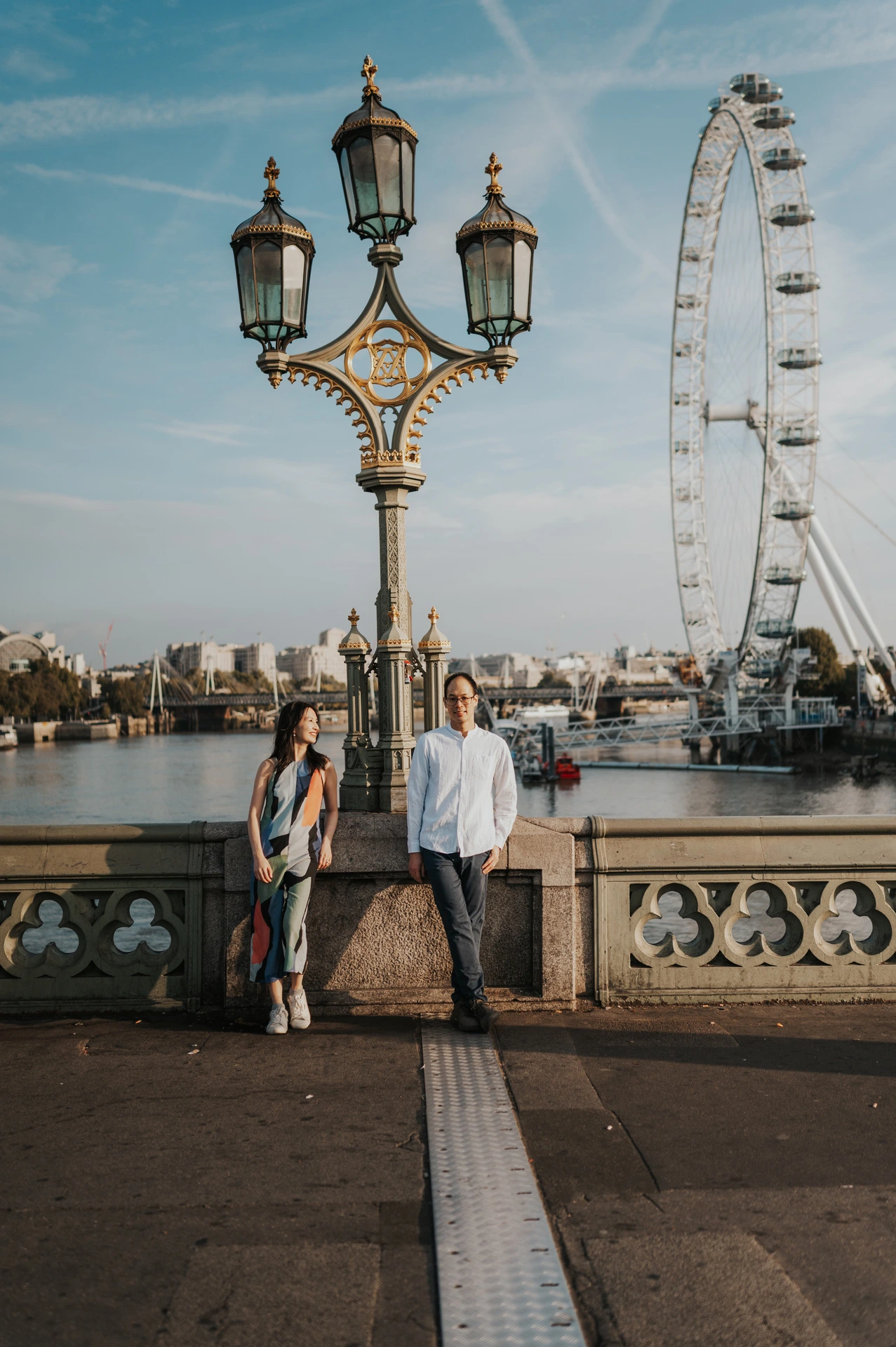 Wide portrait of the couple standing beneath an ornate Westminster Bridge lamp, with the London Eye across the river, during their London couple photoshoot.