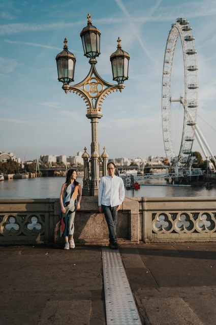 London couple photoshoot beneath the Westminster Bridge lamps Wide portrait of the couple standing beneath an ornate Westminster Bridge lamp, with the London Eye across the river, during their London couple photoshoot.