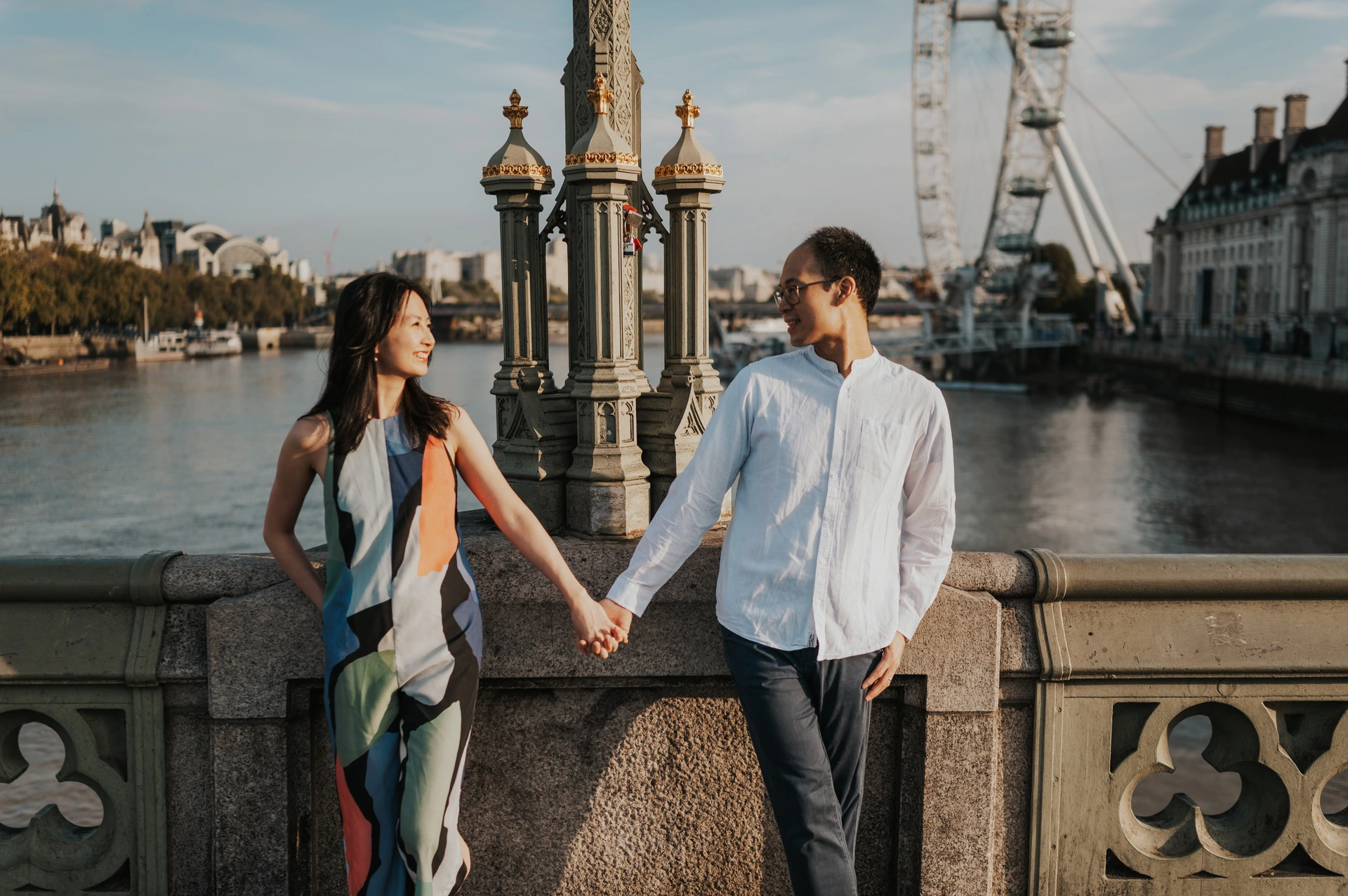 London couple photoshoot holding hands with a London Eye view Couple holding hands beside the stone and green-painted parapet, with the London Eye across the Thames, during their London couple photoshoot.