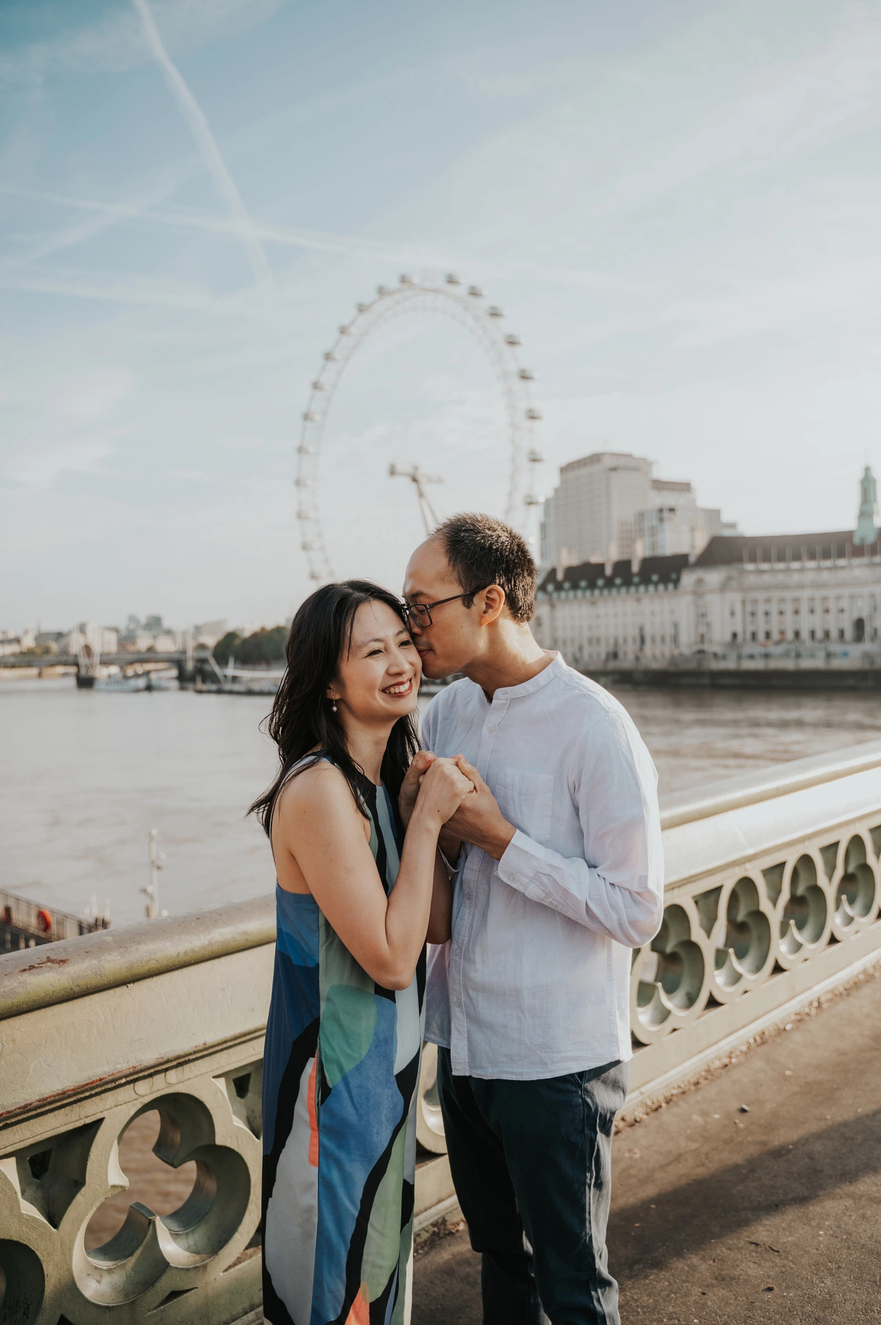Man kissing his partner’s forehead beside Westminster Bridge’s green railing, with the London Eye behind them, during their London couple photoshoot.