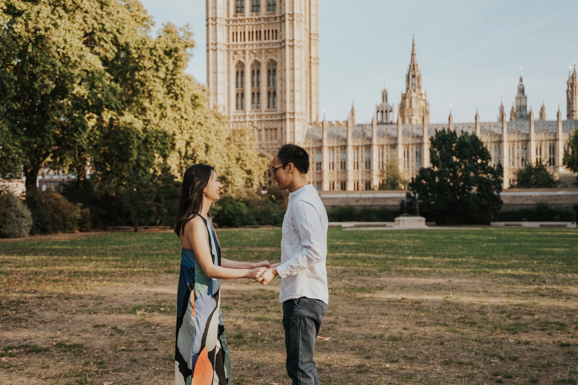 Couple standing face to face and holding hands on the lawn, with Victoria Tower behind them, during their London couple photoshoot.