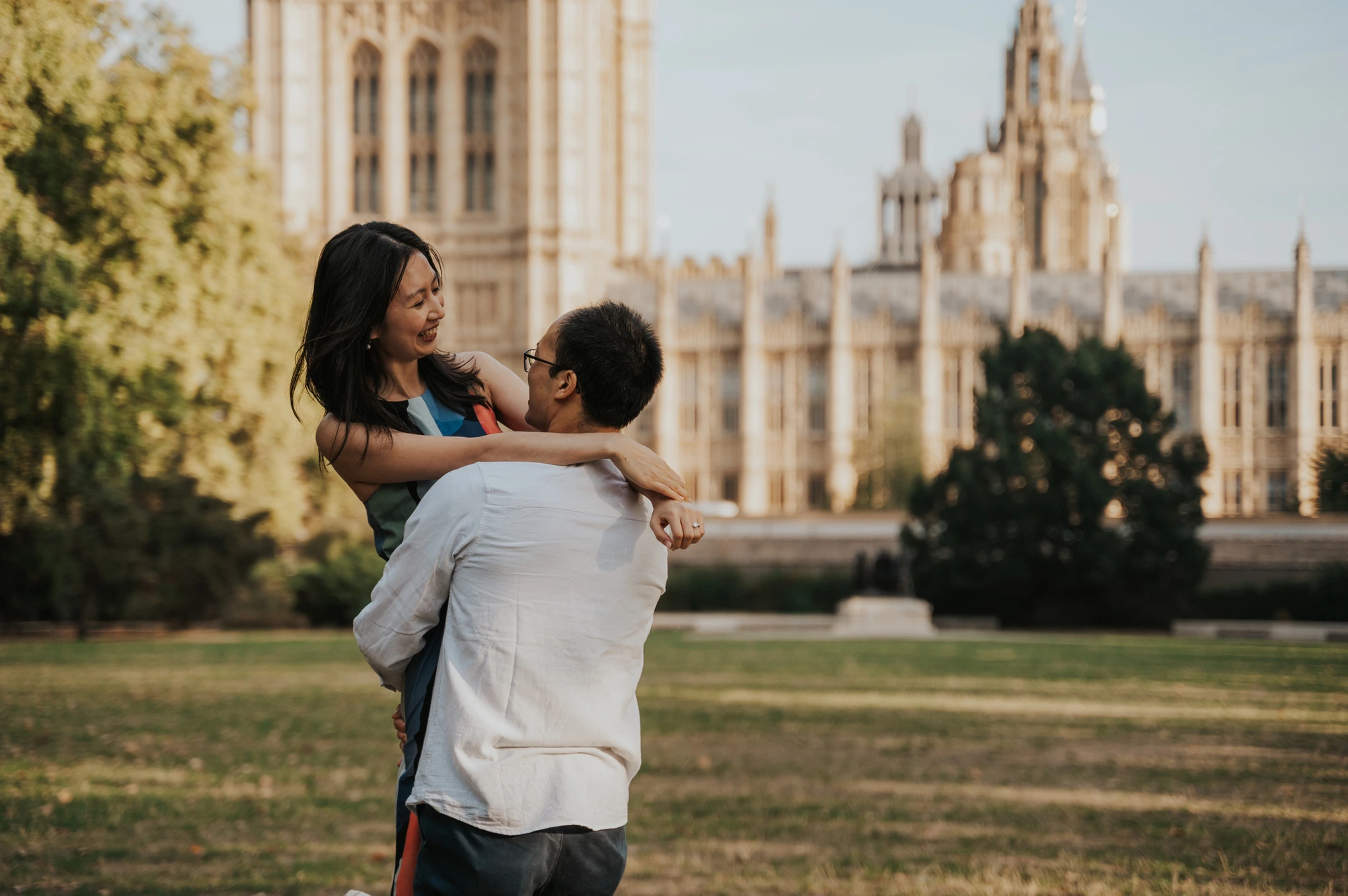 Close portrait of the couple smiling during a lift in Victoria Tower Gardens, with Parliament behind them, during their London couple photoshoot.