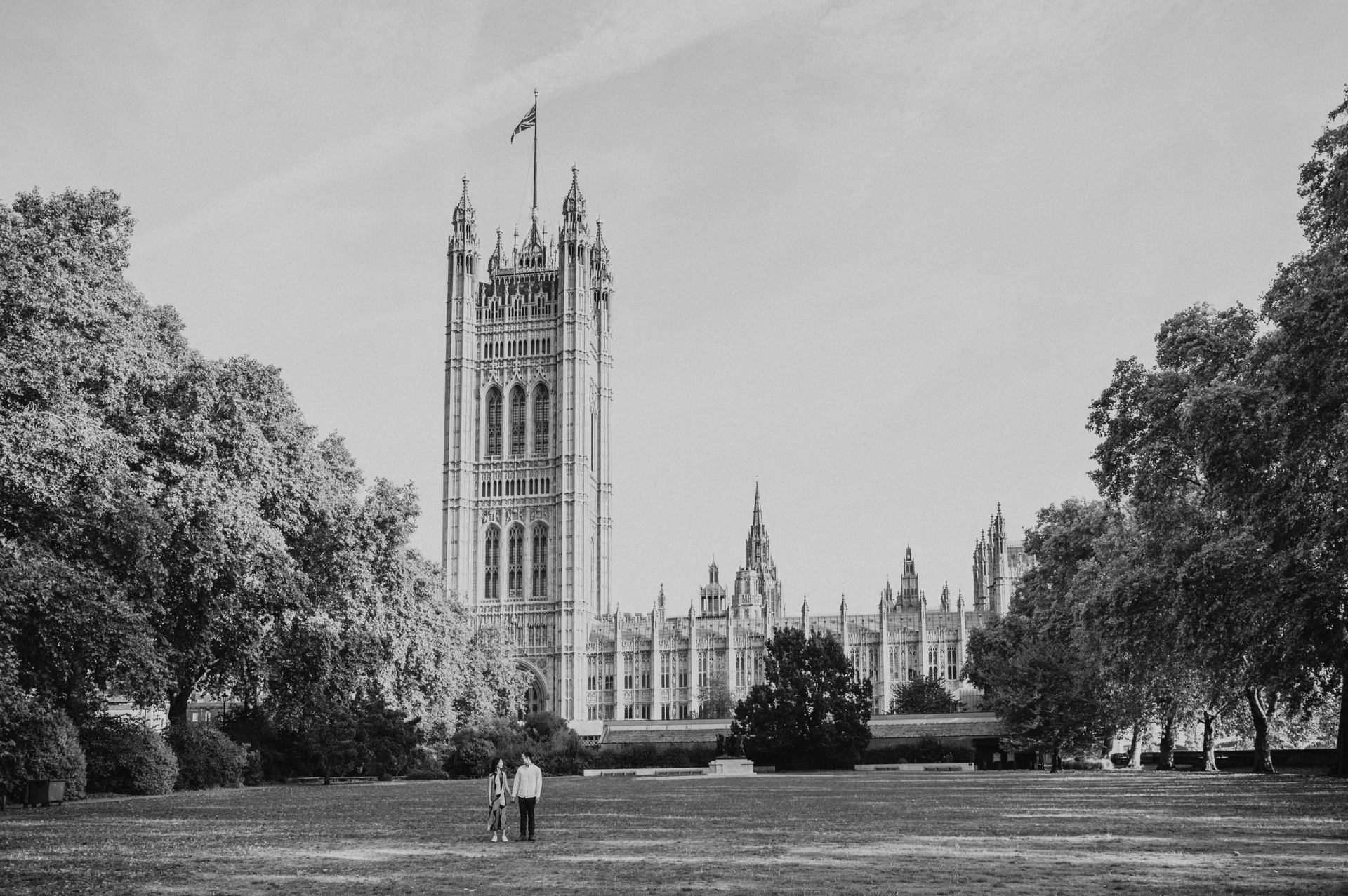 Black-and-white wide view of the couple holding hands on the lawn of Victoria Tower Gardens, with Parliament behind them, from their London couple photoshoot.