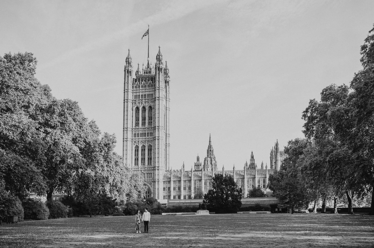 Black-and-white London couple photoshoot across Victoria Tower Gardens Black-and-white wide view of the couple holding hands on the lawn of Victoria Tower Gardens, with Parliament behind them, from their London couple photoshoot.