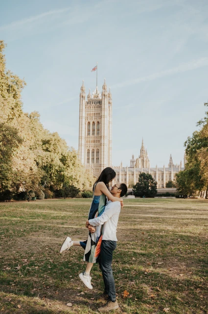 London couple photoshoot with a playful lift in the gardens Wide full-length portrait of the man lifting his partner on the lawn, with Victoria Tower rising behind them, during their London couple photoshoot.