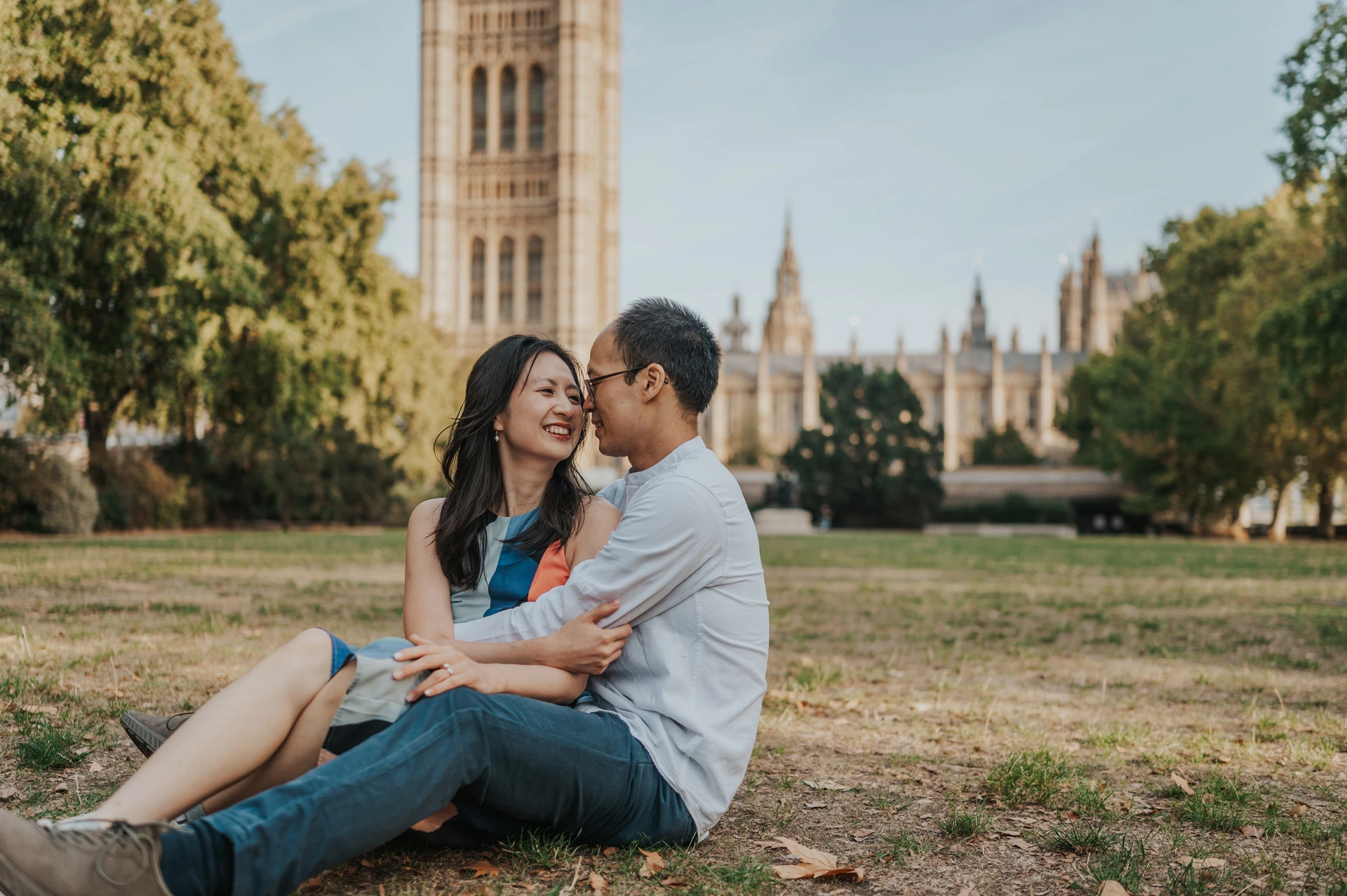 London couple photoshoot smiling on the Westminster garden lawn Close landscape portrait of the couple smiling together while seated on the lawn, with Parliament behind them, during their London couple photoshoot.