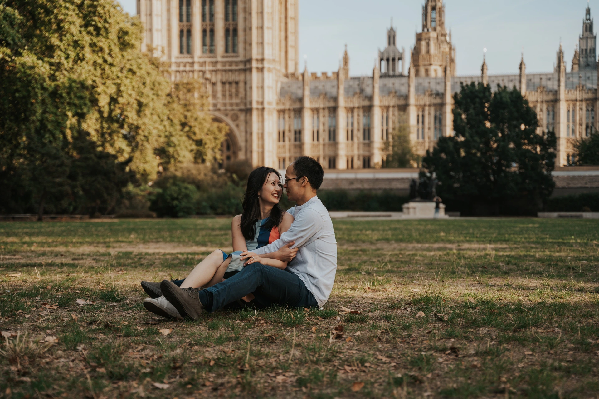 Couple sitting together on the lawn of Victoria Tower Gardens, with Parliament across the background, during their London couple photoshoot.