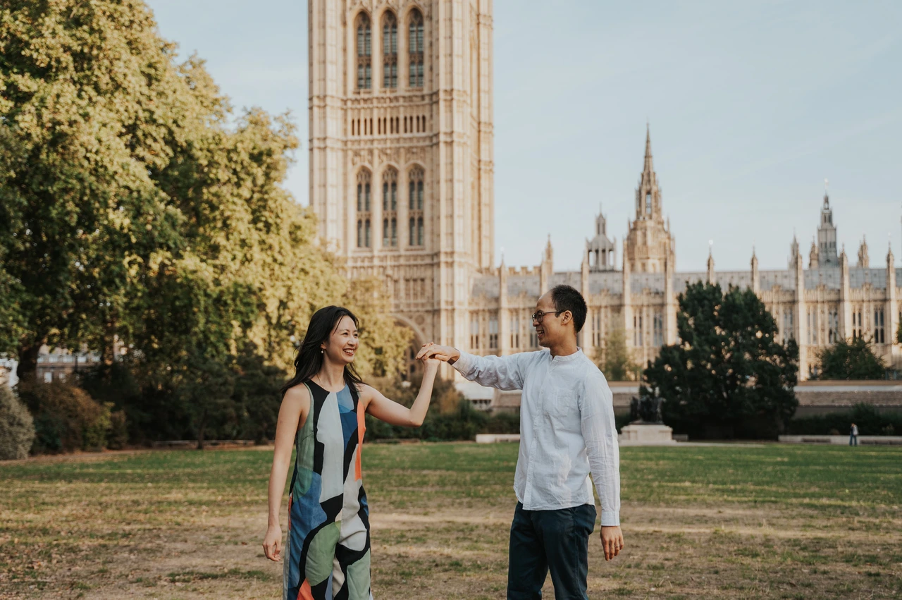 London couple photoshoot turning together beside Parliament Couple turning with their joined hands raised on the lawn, with Parliament behind them, during their London couple photoshoot.