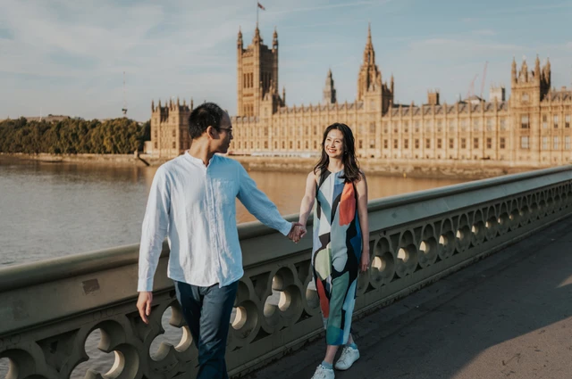 Couple holding hands beside the green parapet of Westminster Bridge, with Parliament across the water, during their London couple photoshoot.