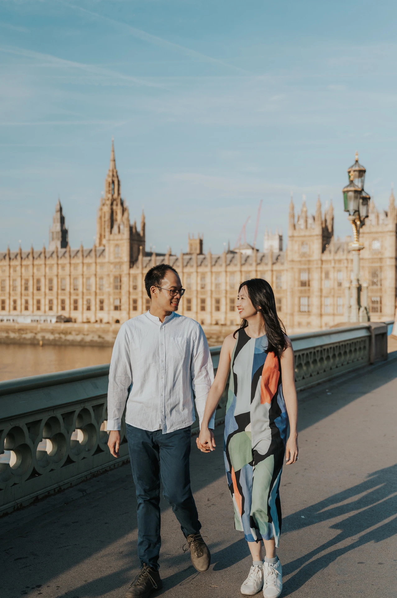 Couple walking hand in hand on Westminster Bridge, with Parliament across the Thames, during their London couple photoshoot.