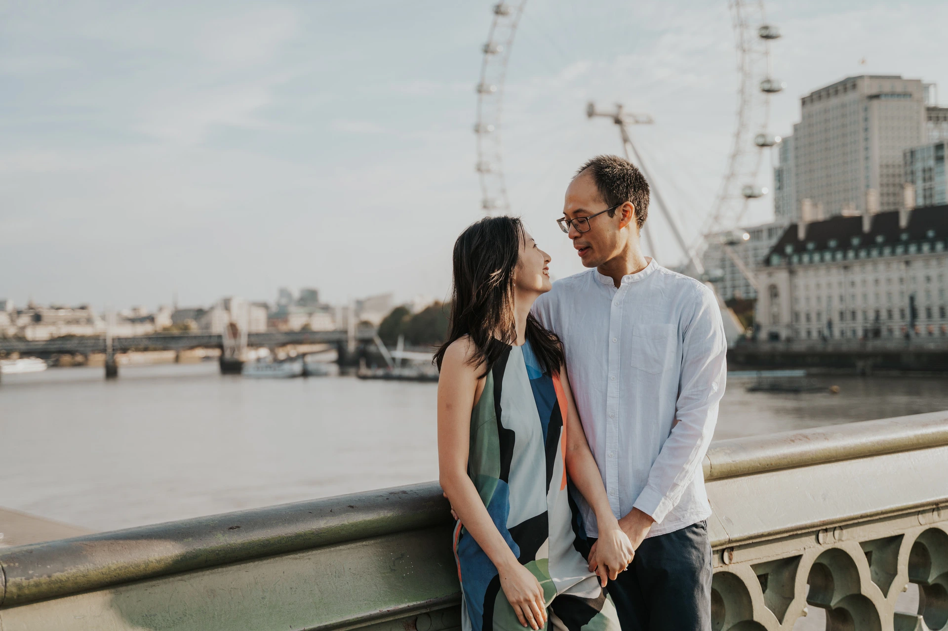 Wide close portrait of the couple looking at each other beside the Thames, with the opposite riverbank behind them, during their London couple photoshoot.