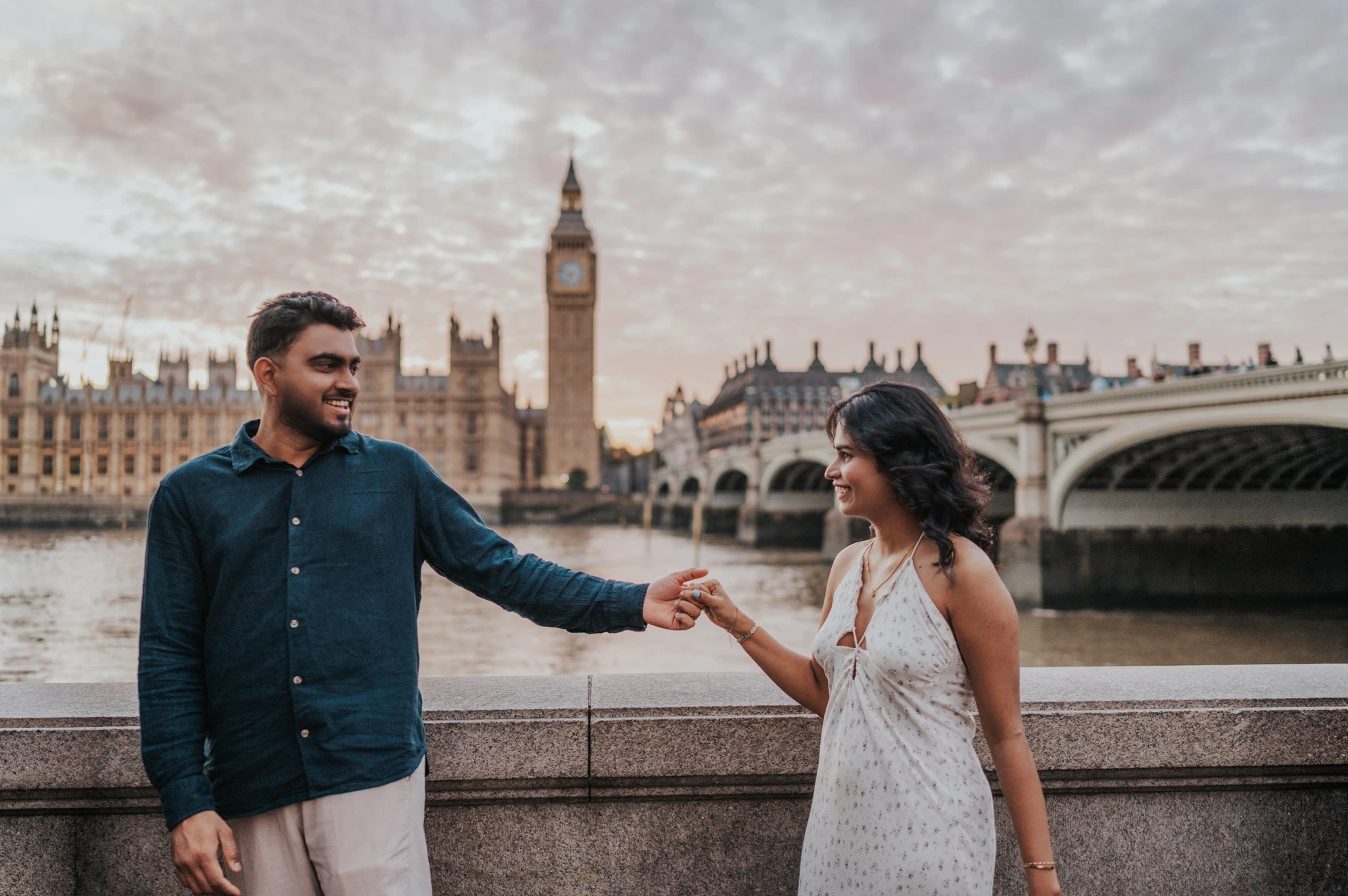London couple photoshoot holding hands with Westminster behind Couple holding one another’s hand across the frame beside the Thames, with Big Ben and Westminster Bridge behind, during their London couple photoshoot.