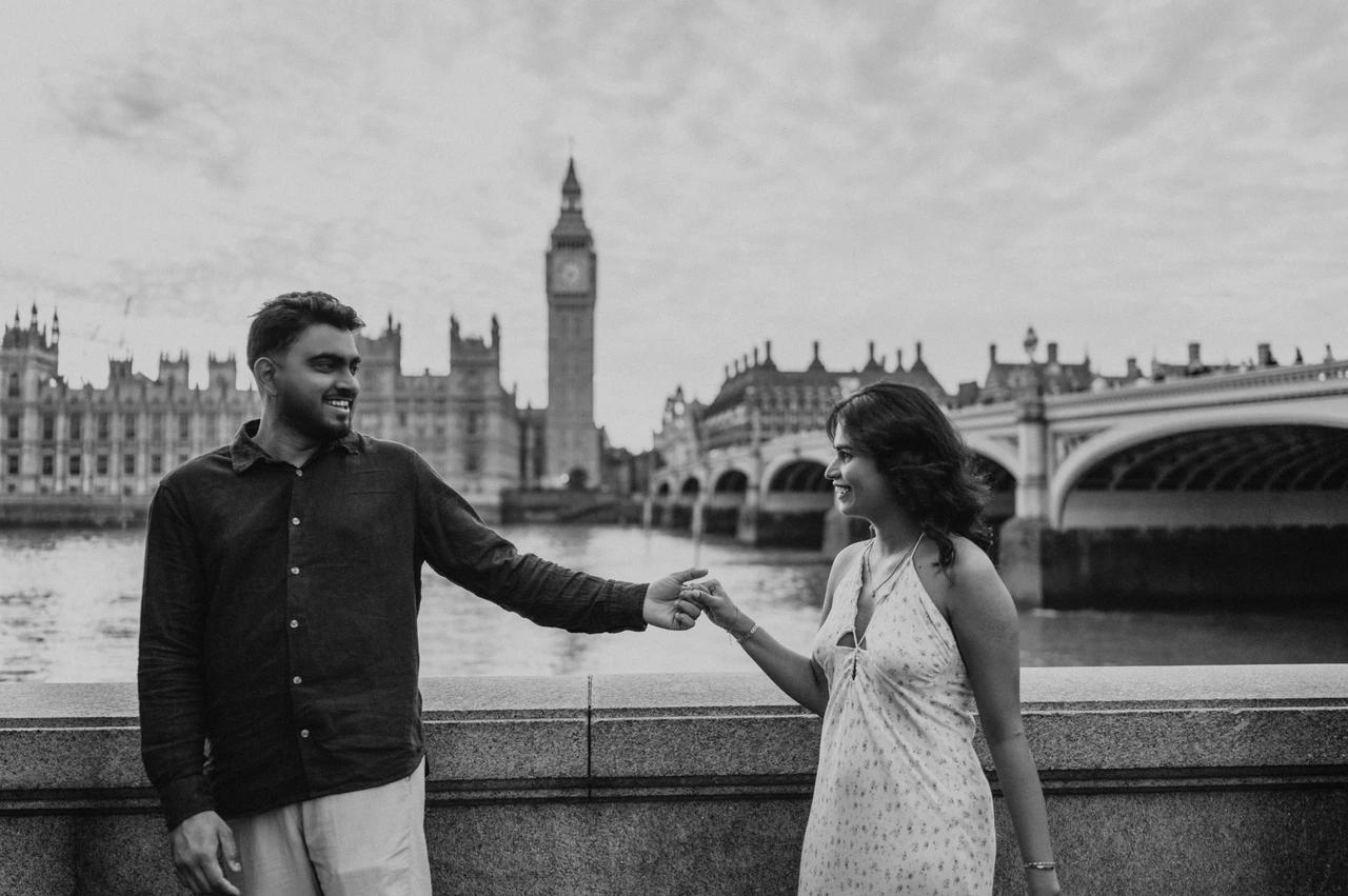 Black-and-white London couple photoshoot with Westminster behind Black-and-white photograph of the couple holding hands beside the Thames, with Big Ben and Westminster Bridge behind, from their London couple photoshoot.