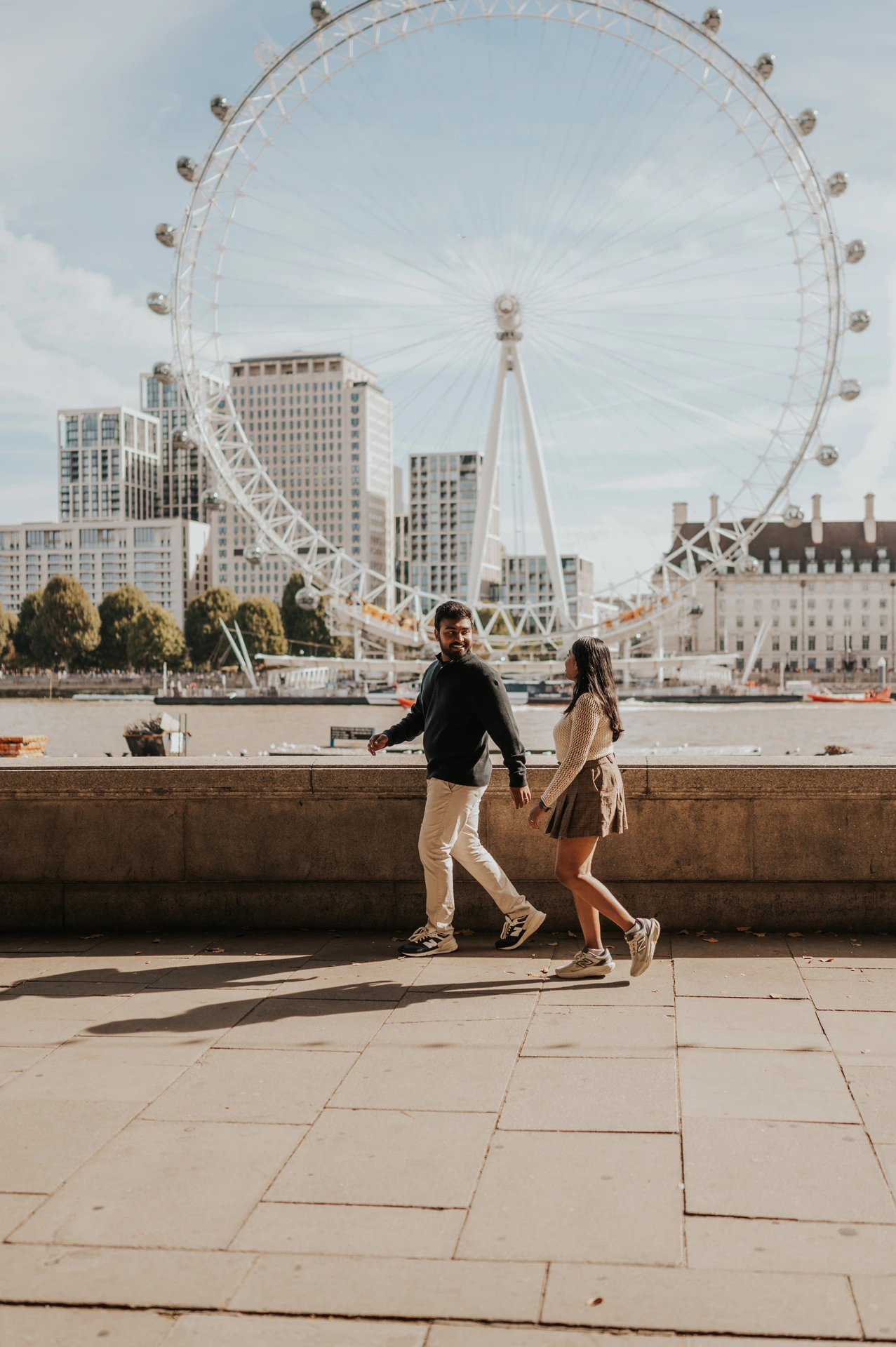 The couple walking beside the stone riverside wall near Westminster Pier, with the full London Eye rising across the Thames behind them during their London couple photoshoot.