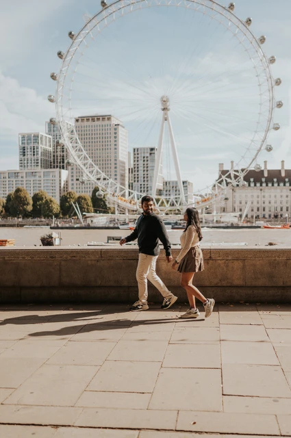 A riverside walk with the London Eye in the background — London couple photoshoot The couple walking beside the stone riverside wall near Westminster Pier, with the full London Eye rising across the Thames behind them during their London couple photoshoot.