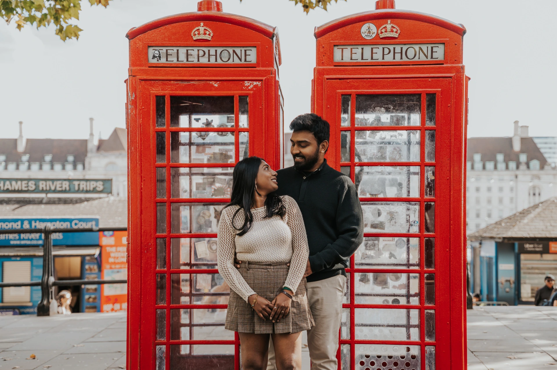The couple standing close in front of two red telephone boxes near Westminster Pier, with the man behind the woman during their London couple photoshoot.