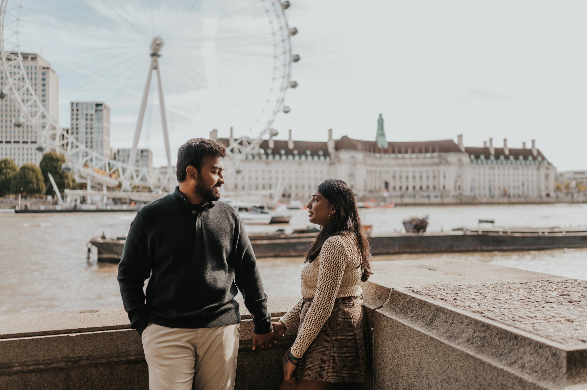 The couple facing each other beside the stone riverside wall near Westminster Pier, with the Thames, County Hall and the London Eye behind them during their London couple photoshoot.