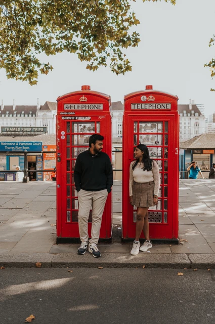 Full-length portraits beside Westminster’s red telephone boxes — London couple photoshoot A full-length portrait of the couple standing beside one another at two red telephone boxes near Westminster Pier, beneath overhanging leaves during their London couple photoshoot.