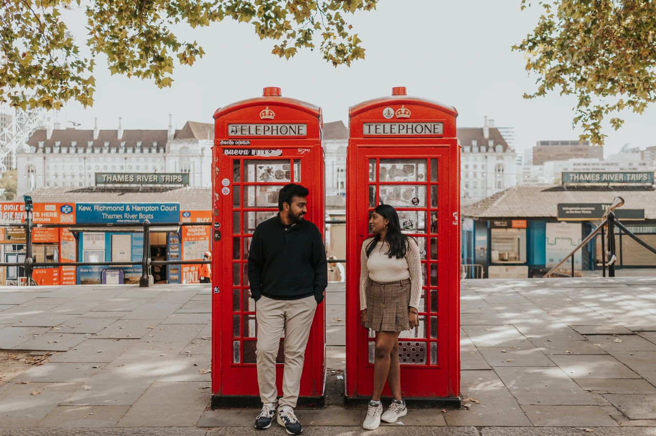 A wide view of the red telephone boxes near Westminster Pier — London couple photoshoot A wide view of the couple looking at each other beside two red telephone boxes near Westminster Pier, with tree branches overhead during their London couple photoshoot.