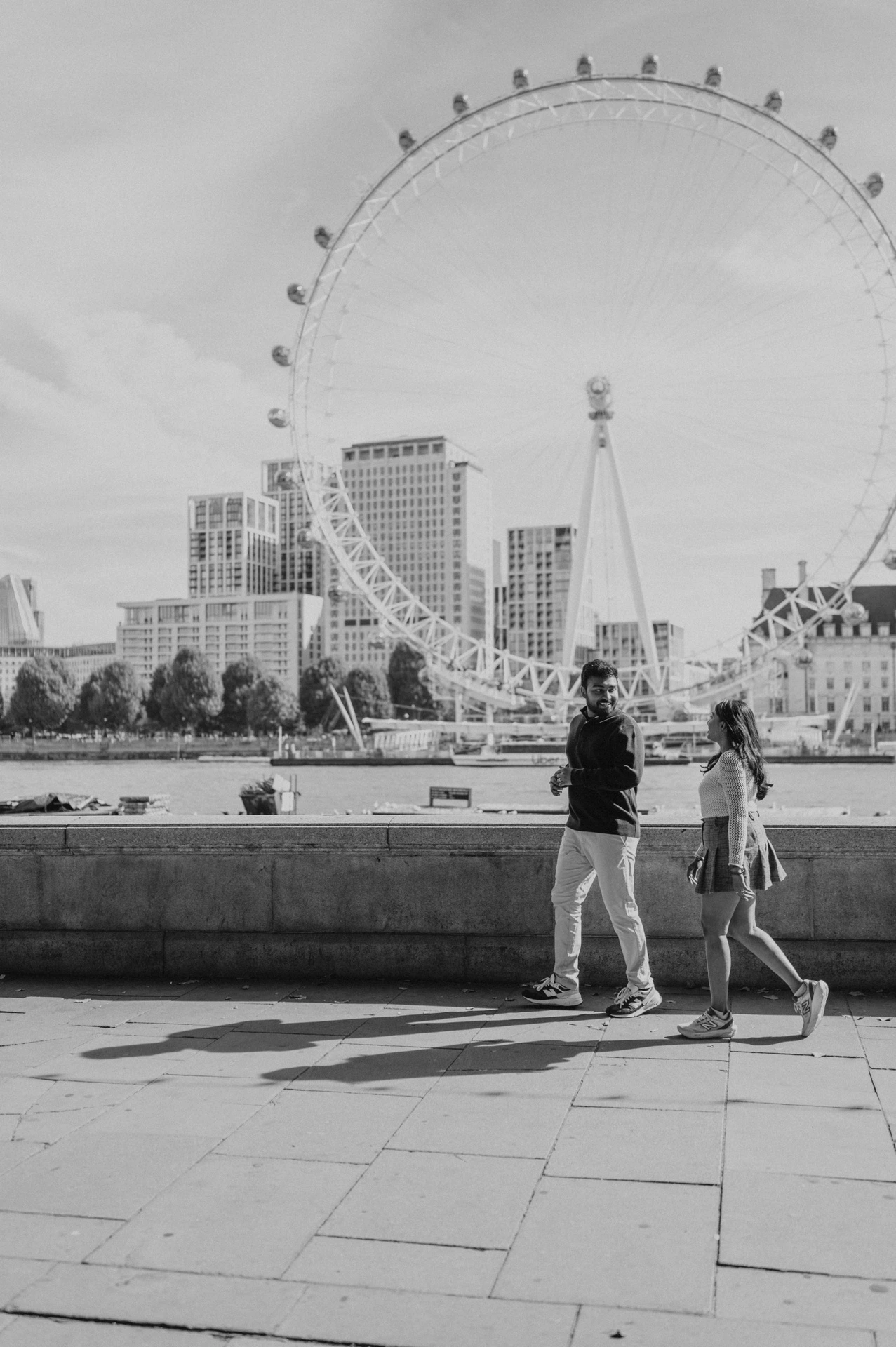 A full-length black-and-white view of the couple walking beside the stone riverside wall near Westminster Pier, with the London Eye across the Thames during their London couple photoshoot.