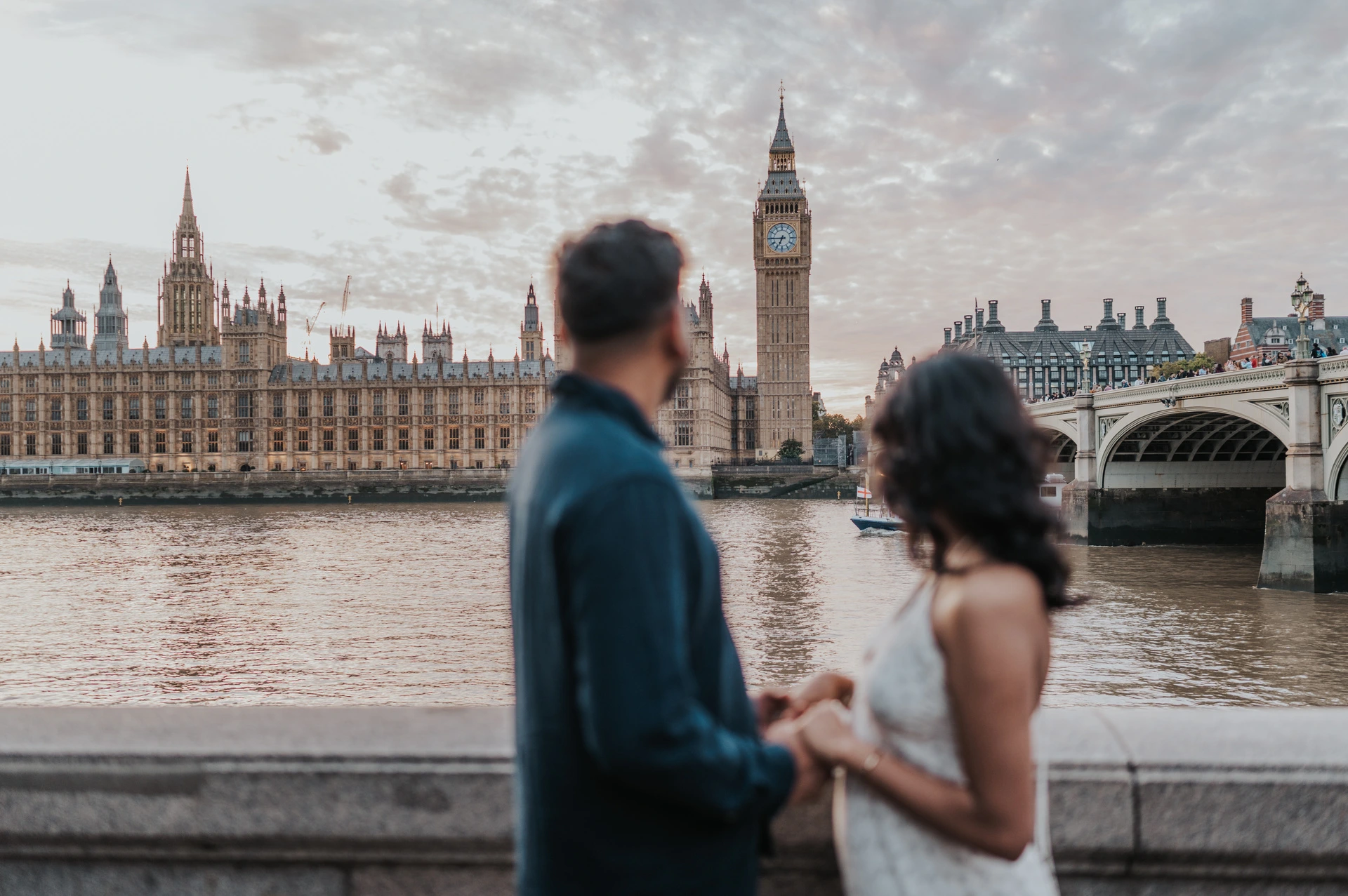 Couple seen from behind beside the Thames, with the Houses of Parliament and Big Ben across the water, during their London couple photoshoot.