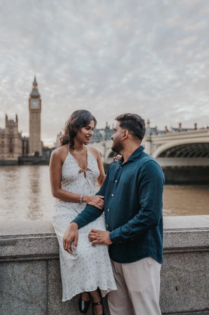 London couple photoshoot beside the riverside wall Woman seated on the riverside wall beside her partner, with Big Ben across the water, during their London couple photoshoot.