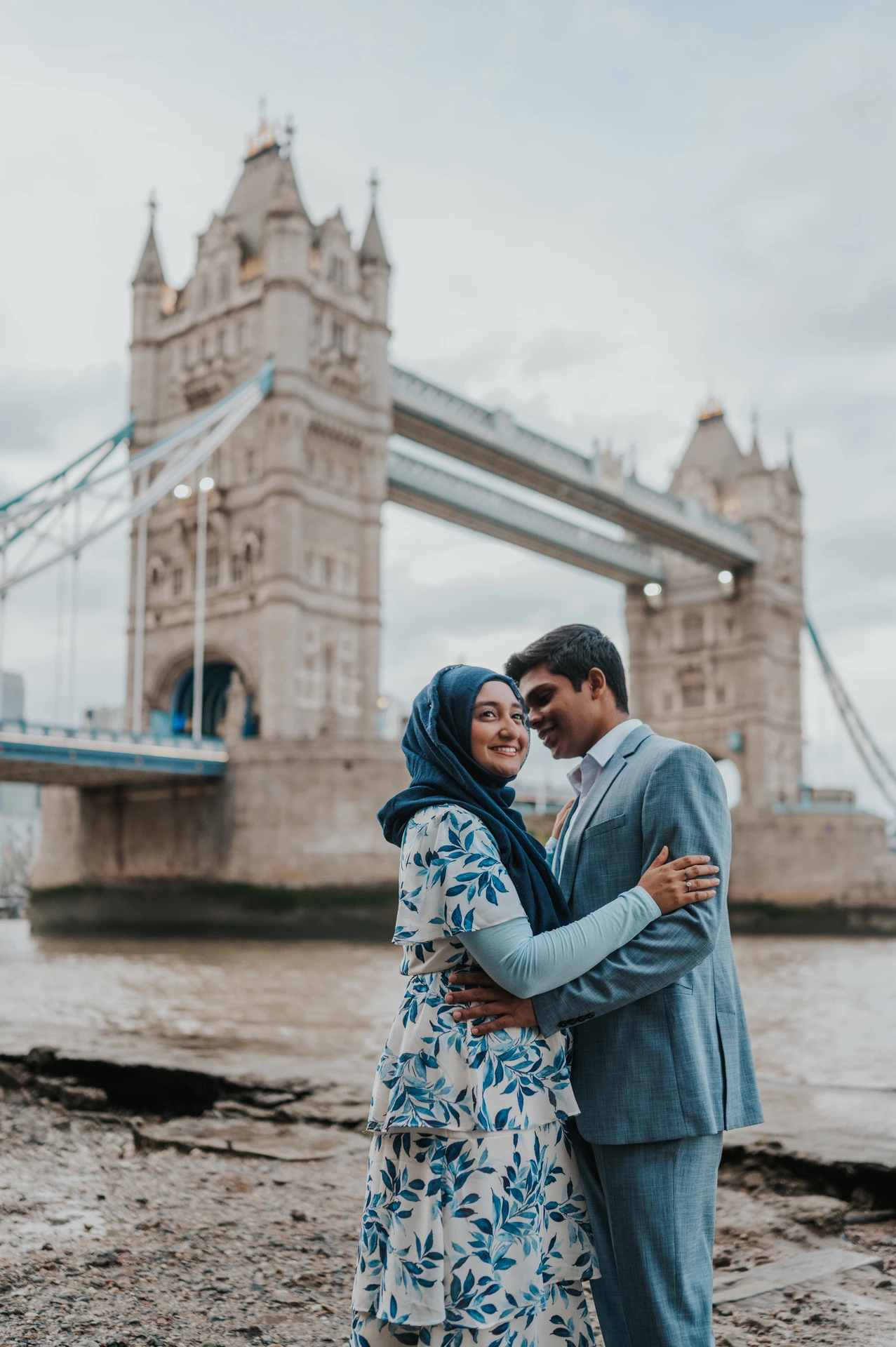 A couple leaning close in an embrace beside the Thames with Tower Bridge in the background during their London engagement photoshoot.
