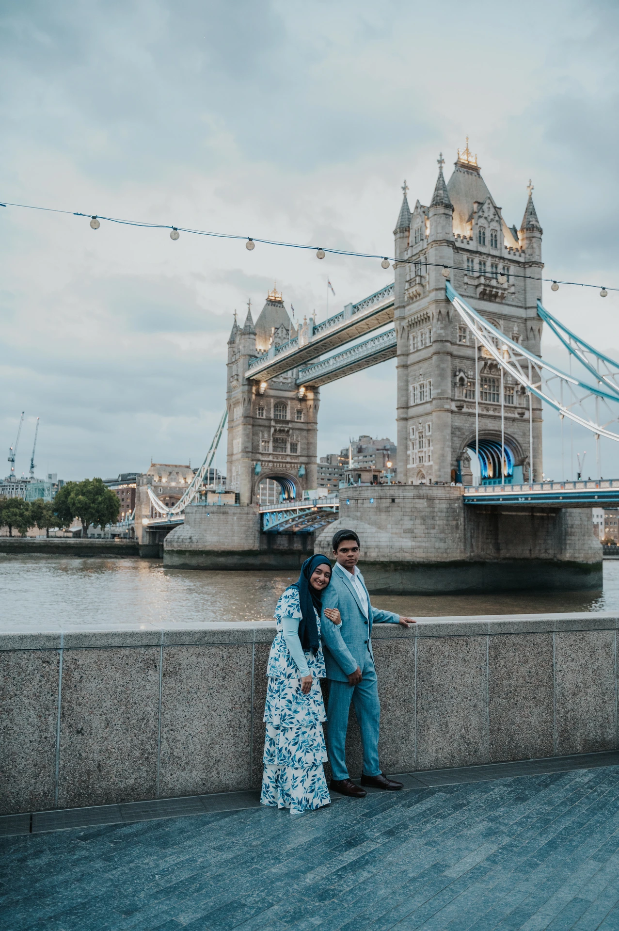 An engaged couple beside a riverside wall with Tower Bridge spanning the Thames behind them during their London engagement photoshoot.