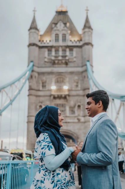 London engagement photoshoot: a portrait with Tower Bridge’s stone tower A couple looking at each other beside Tower Bridge’s blue railing with a stone tower behind during their London engagement photoshoot.