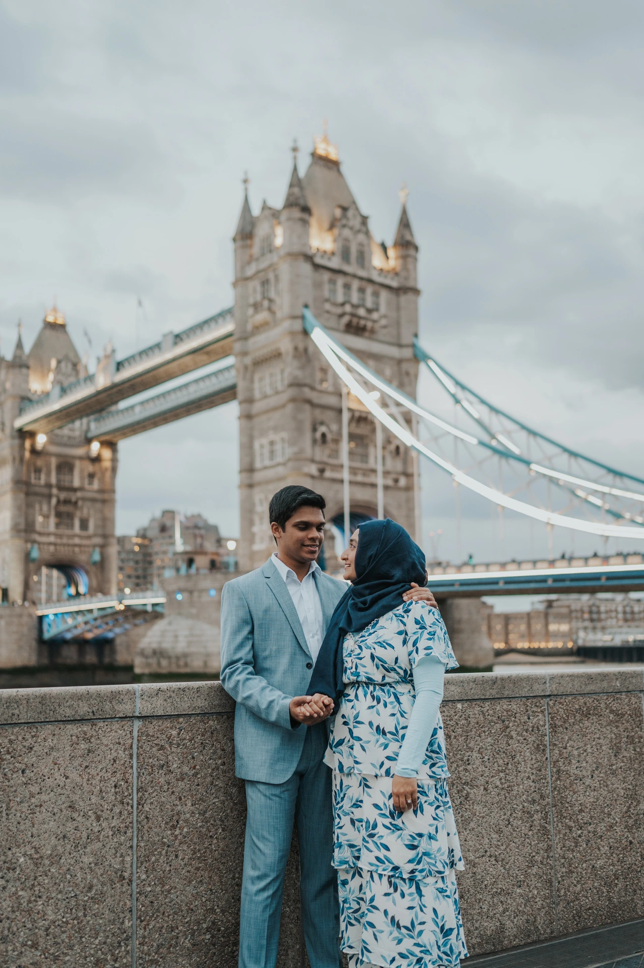 A couple looking at each other beside the riverside wall with Tower Bridge above them during their London engagement photoshoot.