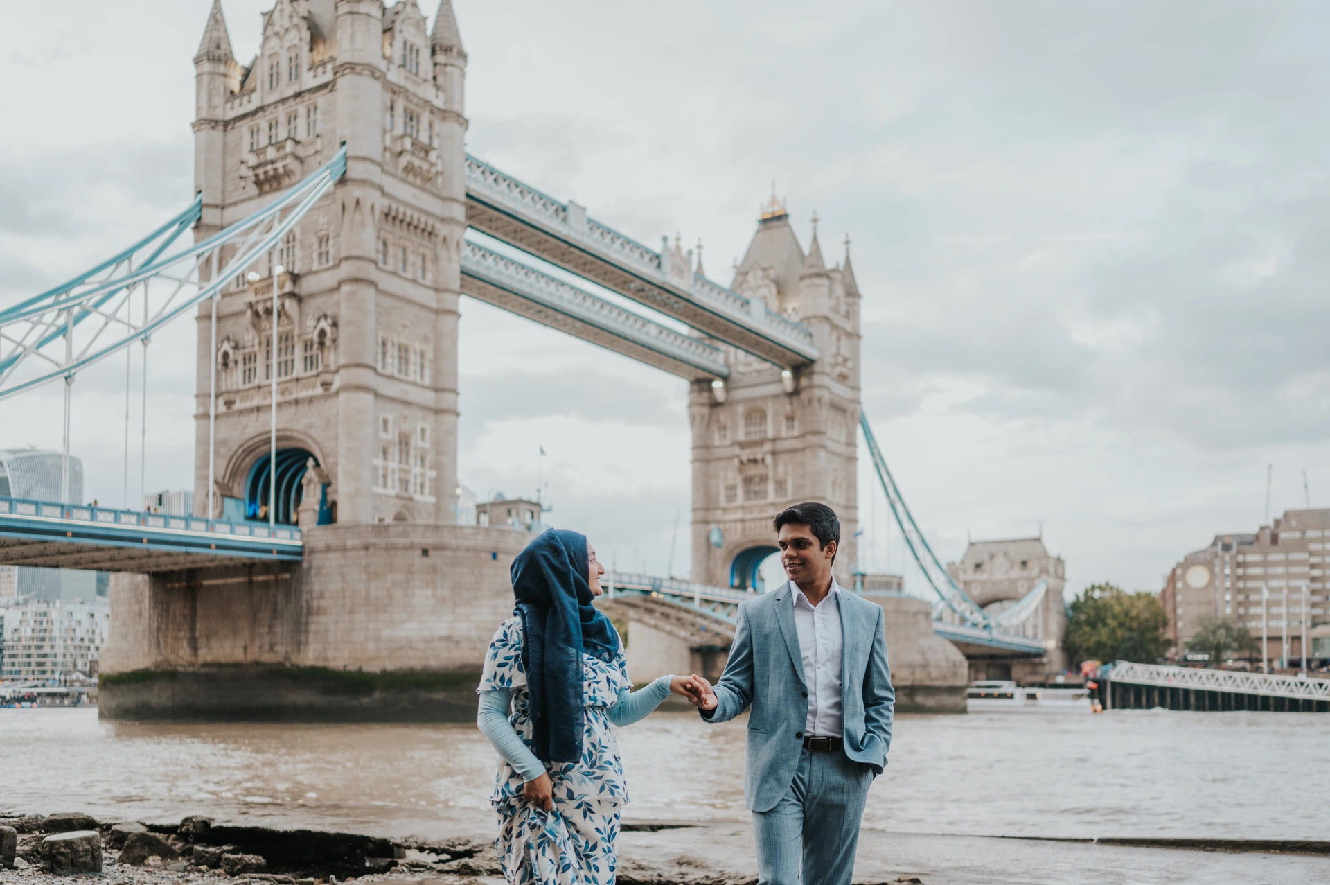 Wide view of a couple walking together with Tower Bridge and the Thames behind them during their London engagement photoshoot.
