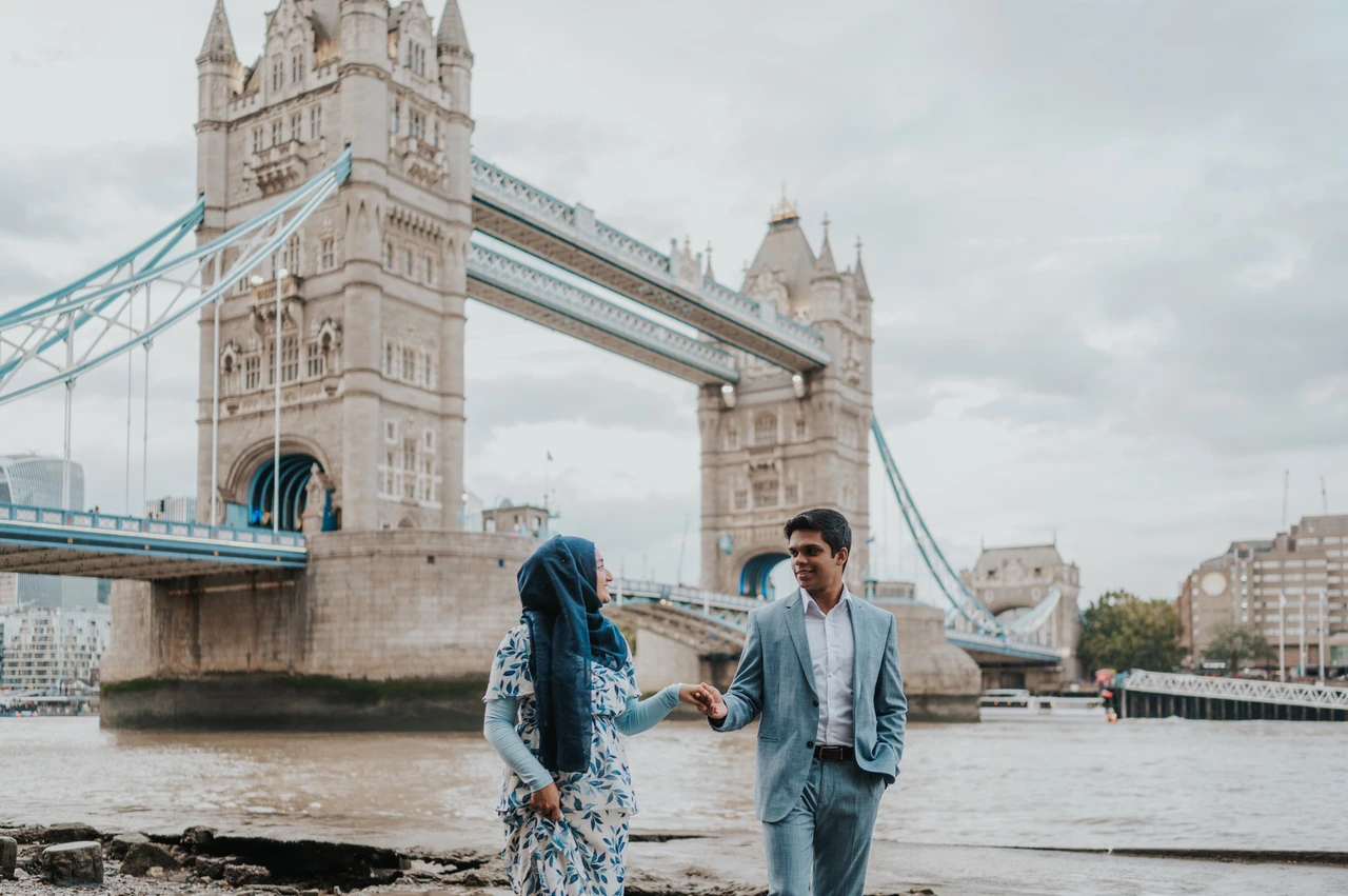 London engagement photoshoot: a riverside walk with Tower Bridge behind Wide view of a couple walking together with Tower Bridge and the Thames behind them during their London engagement photoshoot.