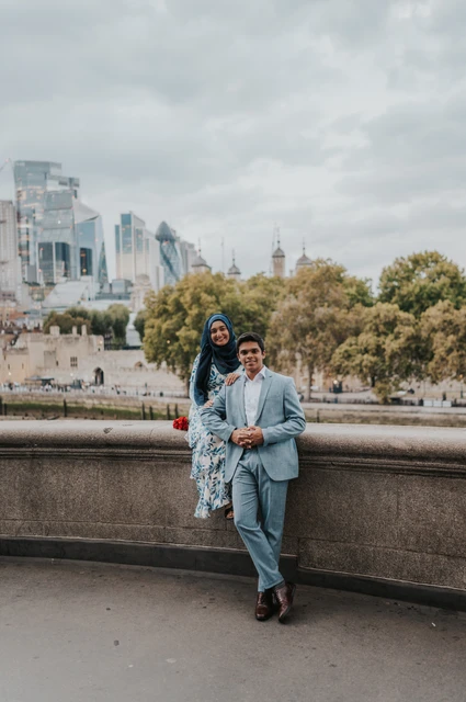 London engagement photoshoot: a seated portrait overlooking the Thames A couple sitting close together on a stone ledge with trees and buildings across the Thames behind them during their London engagement photoshoot.