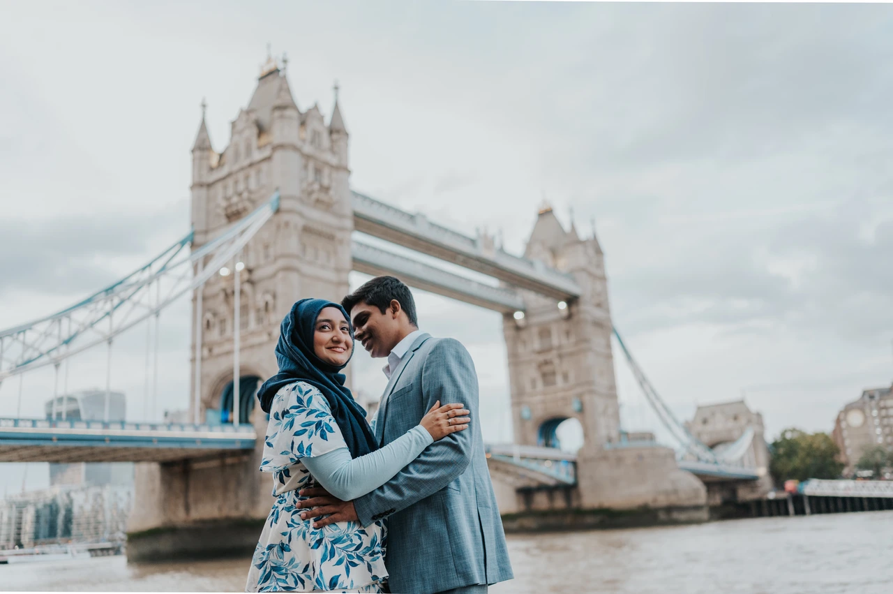 London engagement photoshoot: a smiling portrait with Tower Bridge A woman smiling as her partner leans towards her with Tower Bridge across the Thames behind them during their London engagement photoshoot.