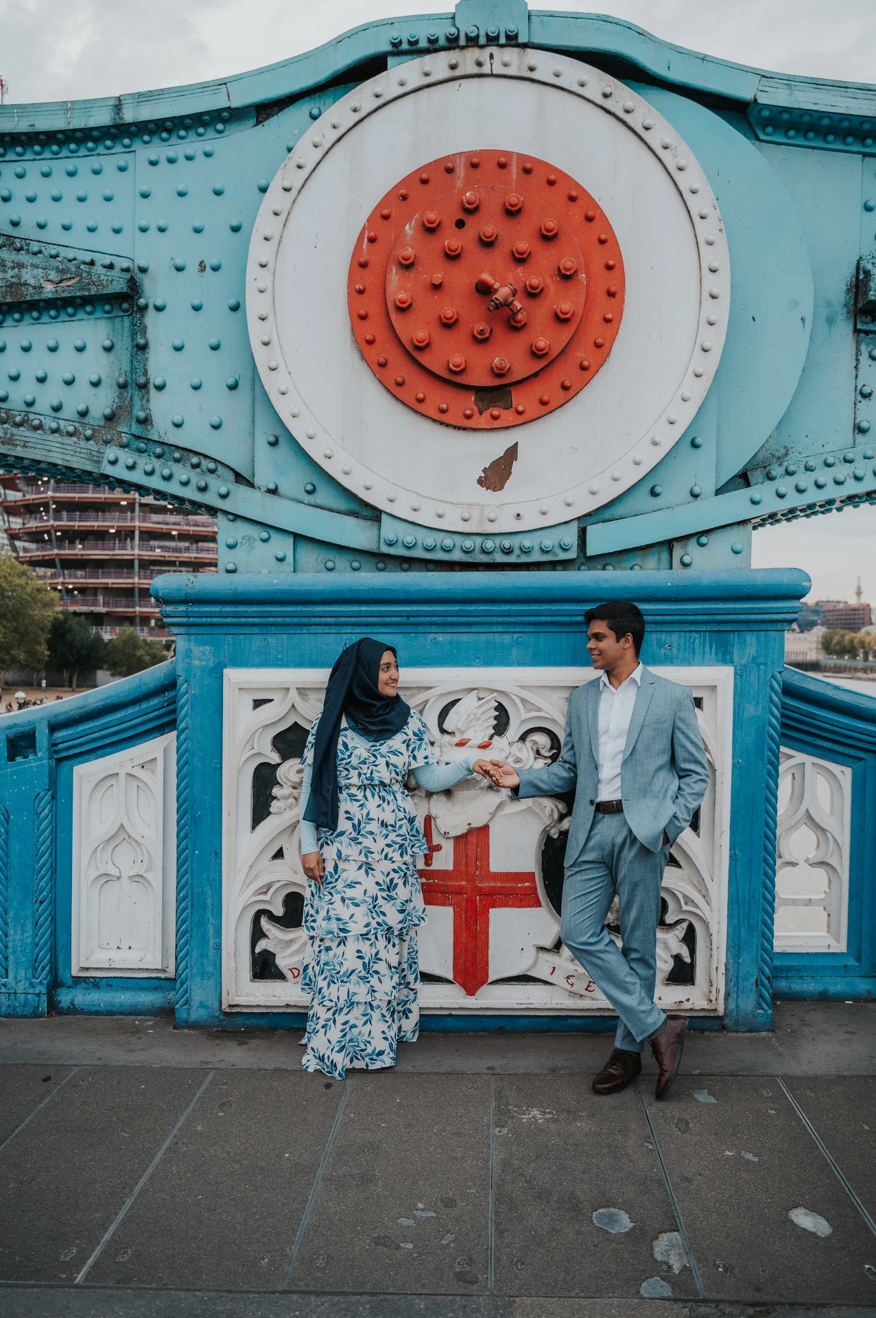 Wide view of a couple beneath Tower Bridge’s blue steelwork and circular red detail during their London engagement photoshoot.