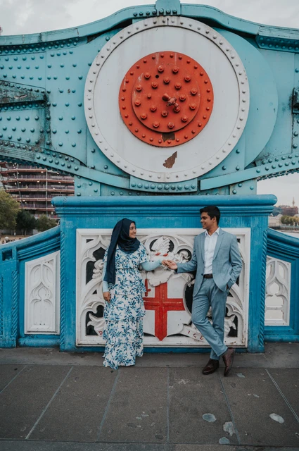 London engagement photoshoot: a wide portrait beneath the bridge steelwork Wide view of a couple beneath Tower Bridge’s blue steelwork and circular red detail during their London engagement photoshoot.
