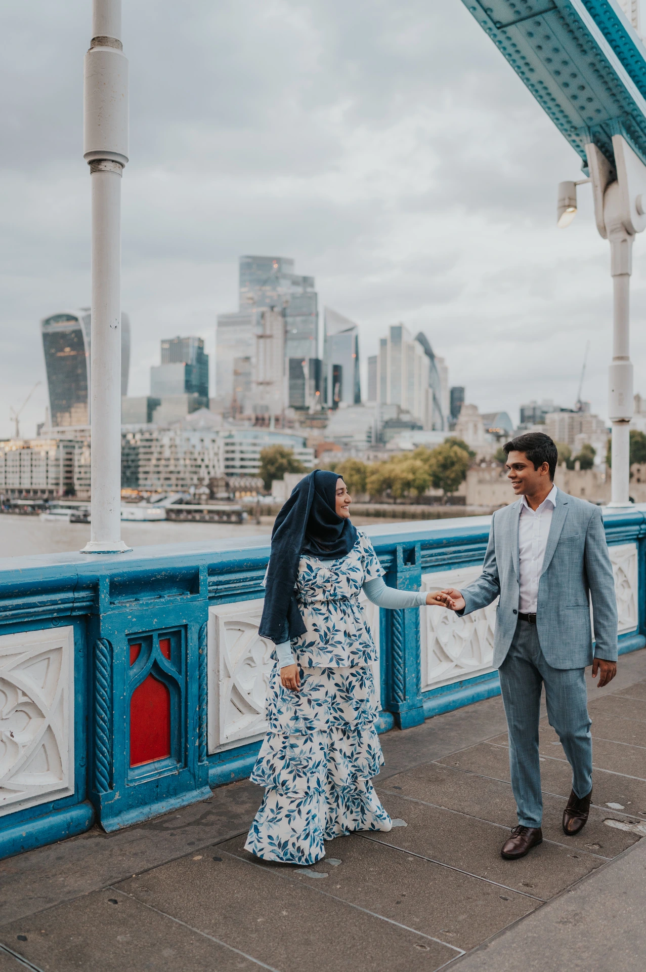 Wide portrait of a couple holding hands on Tower Bridge beside its blue railing and a white lamp post during their London engagement photoshoot.