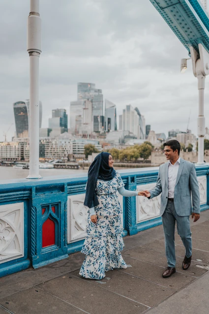 London engagement photoshoot: a wide portrait on Tower Bridge Wide portrait of a couple holding hands on Tower Bridge beside its blue railing and a white lamp post during their London engagement photoshoot.