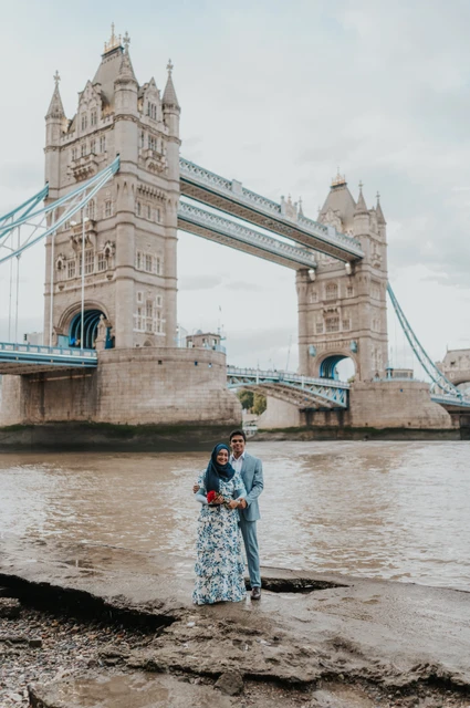 London engagement photoshoot: an embrace beneath the bridge towers A couple embracing below Tower Bridge with the Thames around the foreshore during their London engagement photoshoot.