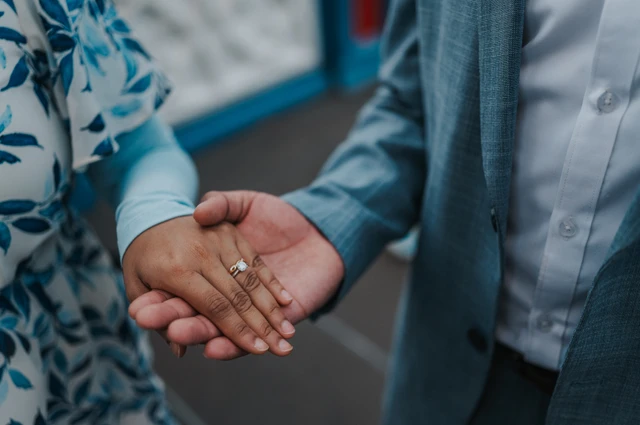 Close-up of an engagement ring on the woman’s hand resting in her partner’s palm beside Tower Bridge’s blue railing during their London engagement photoshoot.