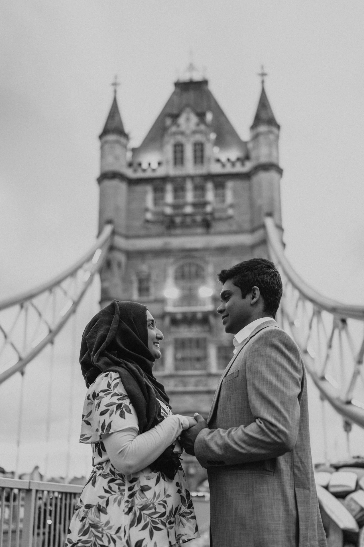 Black-and-white close portrait of a couple beside Tower Bridge’s blue railing and stone tower during their London engagement photoshoot.
