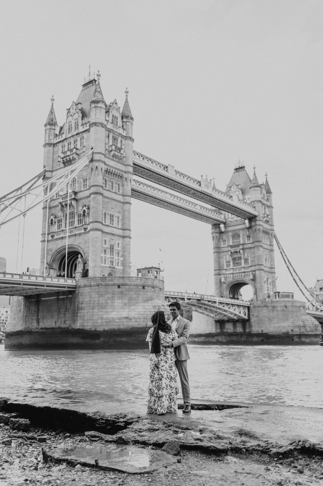 Black-and-white view of a couple embracing on the foreshore below Tower Bridge during their London engagement photoshoot.