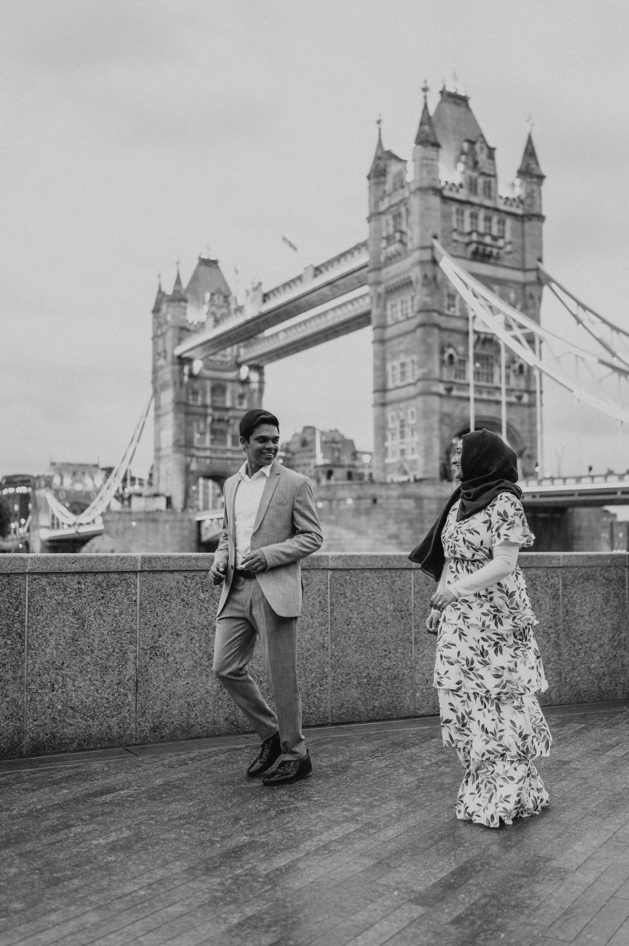 Black-and-white photograph of a couple walking beside a riverside wall with Tower Bridge behind during their London engagement photoshoot.