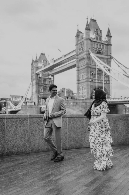 London engagement photoshoot: black-and-white movement along the riverside Black-and-white photograph of a couple walking beside a riverside wall with Tower Bridge behind during their London engagement photoshoot.