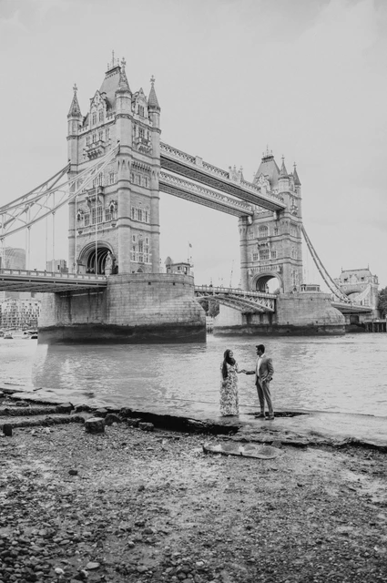 London engagement photoshoot: black-and-white portrait below Tower Bridge Black-and-white wide portrait of a couple on the foreshore beneath Tower Bridge during their London engagement photoshoot.