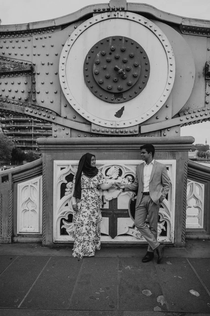 London engagement photoshoot: black-and-white portrait beneath the bridge steelwork Black-and-white portrait of a couple beneath a large circular steel detail on Tower Bridge during their London engagement photoshoot.