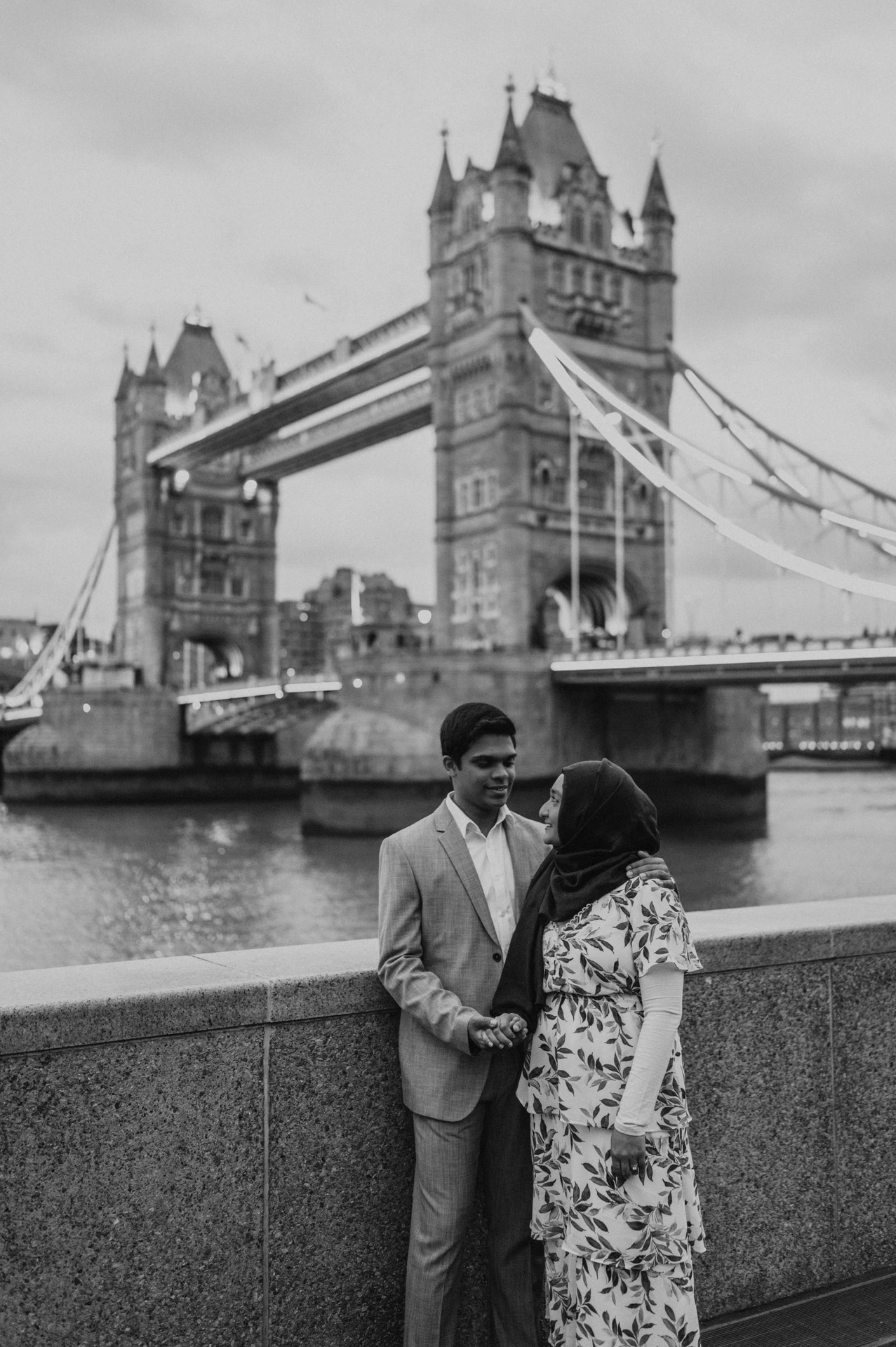 Black-and-white portrait of a couple looking at each other beside the Thames and Tower Bridge during their London engagement photoshoot.