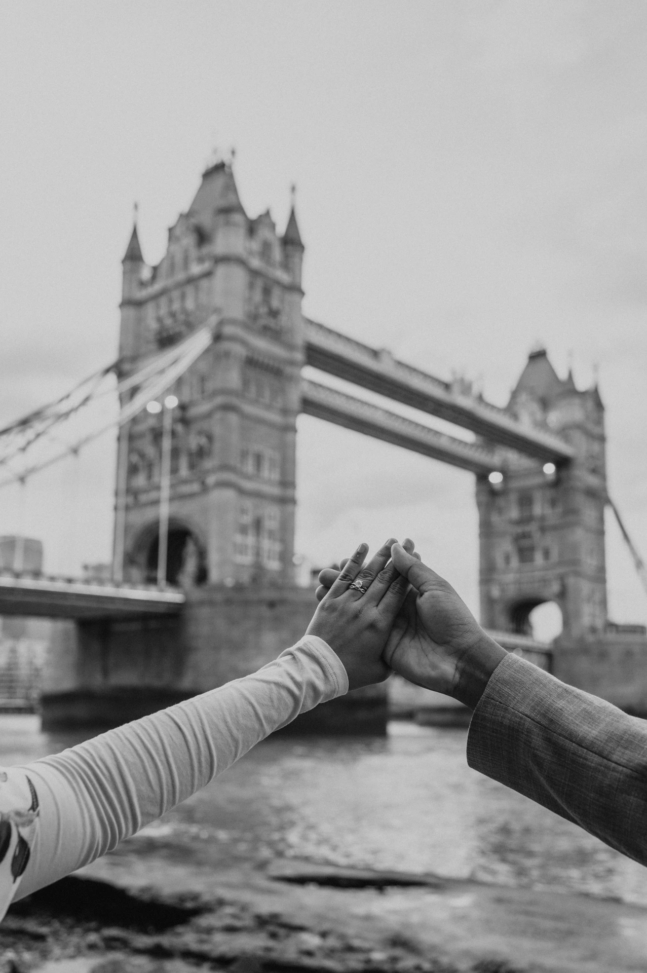 Black-and-white detail of joined hands and an engagement ring with Tower Bridge behind during their London engagement photoshoot.