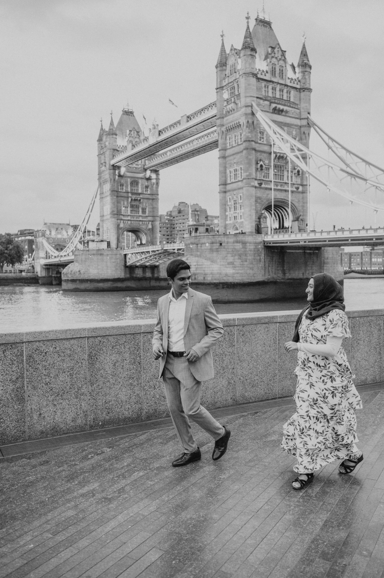 Black-and-white portrait of a couple beside the riverside wall and Tower Bridge during their London engagement photoshoot.