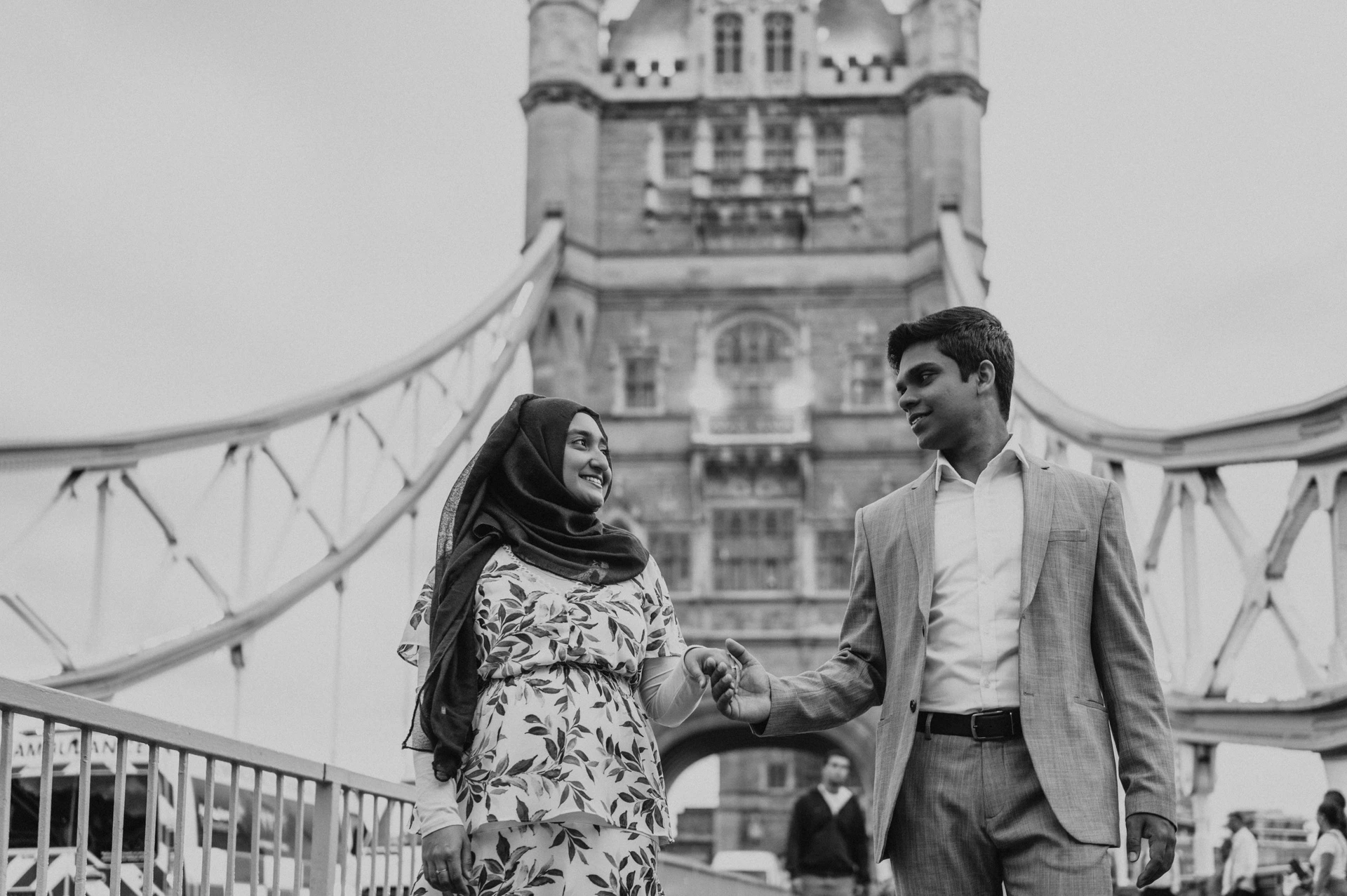 Black-and-white portrait of a couple holding hands beneath Tower Bridge’s suspension details during their London engagement photoshoot.